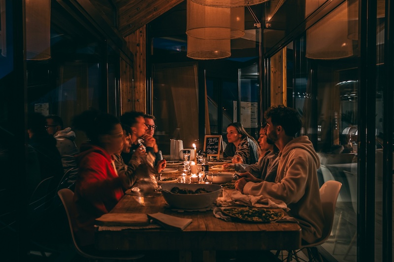 A group of friends shares an intimate candlelit dinner at a long wooden table inside a cozy restaurant with warm ambient light.